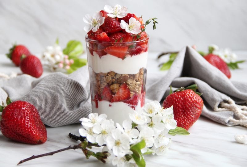 Yogurt parfait with strawberries, flowers and Granola Blues Granola sits on a table surrounded by cloth, strawberries and flowers